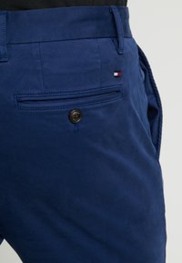 Närbild av blå byxor som visar bakfickan med knapp och en liten röd, vit och marinblå flagga nära midjan.