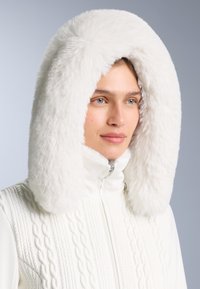 Giacca imbottita bianca con cappuccio in morbida pelliccia sintetica e texture a trecce sul davanti. Il design include una chiusura con zip per funzionalità.