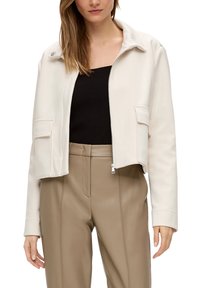 Korte beige jas met een kraag, voorrits en twee borstzakken, gemaakt van zachte stof met een gladde textuur.