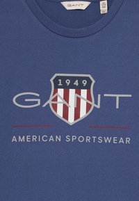 Blå bomull t-skjorte med et skjoldlogo med røde og hvite striper, "GANT" i grått, og teksten "AMERICAN SPORTSWEAR" under.