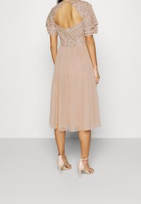 Vista trasera de una mujer que lleva un vestido rosa hasta la rodilla con parte superior de lentejuelas florales, mangas transparentes, espalda descubierta y sandalias de tacón nude.