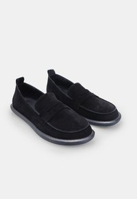 O pereche de mocasini negri din suede, cu talpă din cauciuc și o curea peste partea de sus, expuși pe un fundal gri deschis.