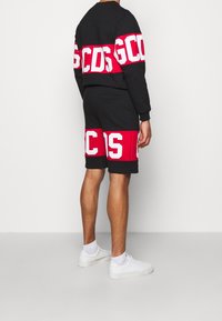Homme portant un sweat-shirt GCDS noir et rouge et un short assorti avec des baskets blanches, debout devant un fond clair uni.
