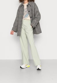 Veste oversize à carreaux noir et blanc, t-shirt graphique crème, pantalon large vert clair, baskets blanches Nike avec des accents néon.