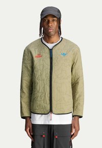 Khaki pikowana kurtka z konturowymi wzorami, czarnym zamkiem, niebieskim logo, czerwonym tekstem i okrągłym dekoltem; nałożona na białą koszulę i ciemne spodnie.