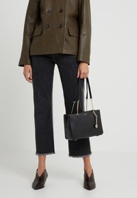 Svart faux läderhandväska med kedjedetaljer och en charm. Slät yta. Bärs med en olivgrön dubbelknäppt jacka och mörka jeans.