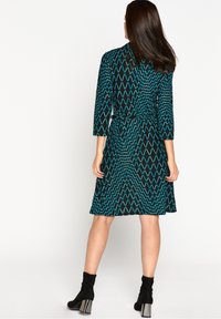 Svart klänning med ett turkos geometriskt mönster, trekvartsärmar och midjeband; knälång och gjord av lättviktsmaterial.