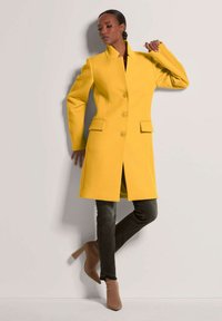 Geel wollen jas met een gestructureerd ontwerp, voorzien van een hoge kraag, drie knopen en twee voorzakken. In combinatie met donkere jeans en beige hakken.