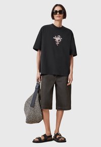 T-shirt nera oversize con stampa floreale e logo, abbinata a pantaloni corti marrone scuro, sandali neri e una borsa a tracolla con motivo.