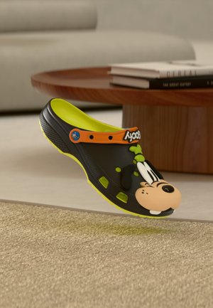 Zoccolo nero e verde neon con il volto del personaggio Goofy sulla punta, un cinturino arancione con la scritta "Goofy", poggiato su un tappeto beige vicino a un tavolo di legno.