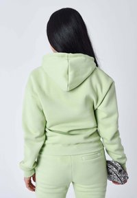 Sudadera verde claro hecha de un material suave con un corte relajado, que cuenta con capucha, puños acanalados y una parte posterior sin costuras. Sosteniendo un bolso clutch gris texturizado.