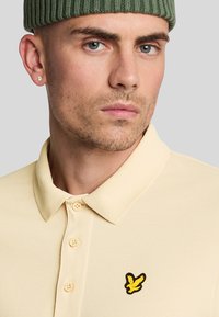 Lichtgeel polo-shirt van textuurstof, met een drie-knopen sluiting en een zwart-geel geborduurd logo op de linkerborst.