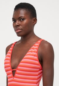 Maillot de bain rayé rouge et rose avec un décolleté en V profond et des bretelles fines. Tissu texturé avec un motif visible. Encolure arrondie et design ajusté.