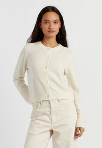 Cardigan color crema realizzato in materiale morbido, con scollo rotondo, chiusura a bottoni e polsini e orlo a coste. Indossato sopra pantaloni di colore chiaro.