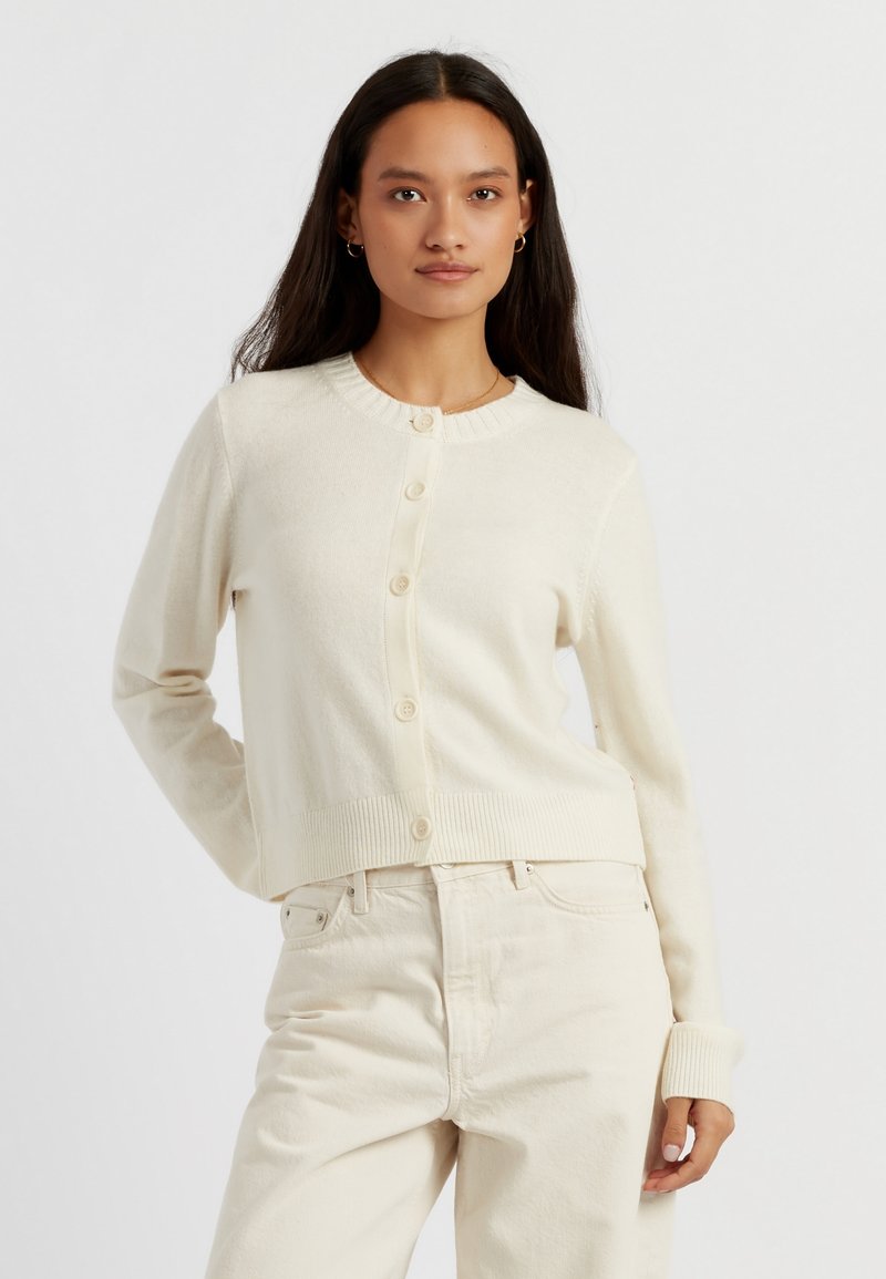 Cardigan color crema realizzato in materiale morbido, con scollo rotondo, chiusura a bottoni e polsini e orlo a coste. Indossato sopra pantaloni di colore chiaro.