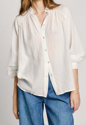 Mujer con blusa blanca suelta abotonada con mangas abullonadas y vaqueros azules, mostrada desde los hombros hasta las caderas sobre fondo liso.