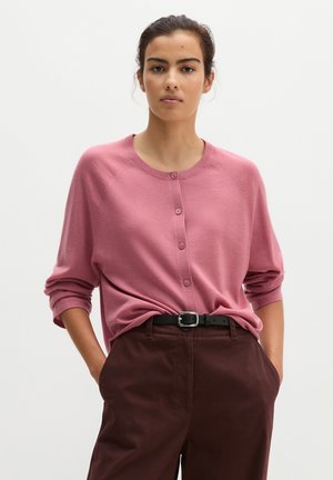 Jeune femme aux cheveux attachés portant un pull rose à boutons et un pantalon marron foncé avec une ceinture noire, mains dans les poches, fond neutre.