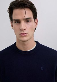 Maglione a maglia blu navy con scollatura a girocollo a coste e un piccolo logo a teschio sul lato sinistro del petto. Tessuto liscio e design aderente.