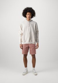 Helle beige Kapuzenjacke mit einer frontalen Tasche und Reißverschluss, kombiniert mit rosa Shorts und weißen Sneakern. Einfaches Design und weicher Stoff.