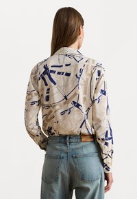 Chemise à manches longues en tissu beige avec des motifs abstraits bleus et bruns. Associée à un jean en denim délavé, offrant un design décontracté.