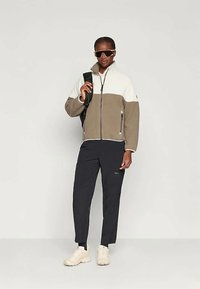 Casaco de fleece com fecho em creme e bege, com bolsos laterais, combinado com calças pretas e ténis bege, complementado com óculos de sol e uma mochila.