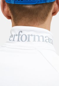 Hvid skjorte med krave, der viser et gråt "performant" logo på bagsiden af kraven. Kraven har en glat tekstur og rene syninger.