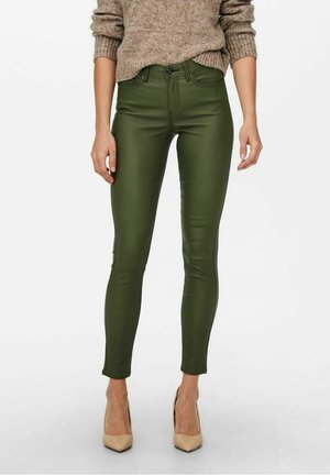 Kobieta ubrana w skórzane, oliwkowozielone, wysokowystające rurki, zestawione z beżowymi wysokimi obcasami i beżowym dzianinowym swetrem z podwiniętymi rękawami.