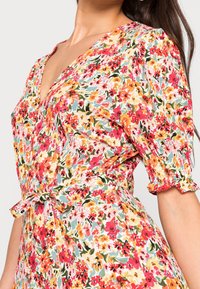 Donna che indossa un vestito floreale con fiori rossi, arancioni e gialli, maniche a sbuffo con polsini elasticizzati e una cintura annodata in vita.