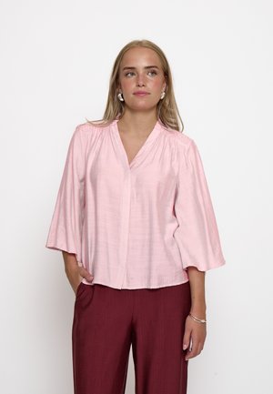 Blonde Frau, die eine hellrosa Bluse mit V-Ausschnitt und weiten Ärmeln sowie dunkelrote Hose trägt, steht mit einer Hand in der Tasche vor weißem Hintergrund.