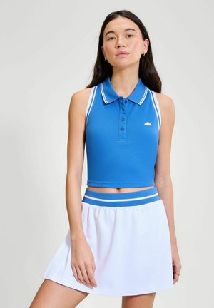 Vrouw die een blauwe mouwloze polo crop top en een witte rok met een blauwe gestreepte tailleband draagt, poserend tegen een effen lichte achtergrond.