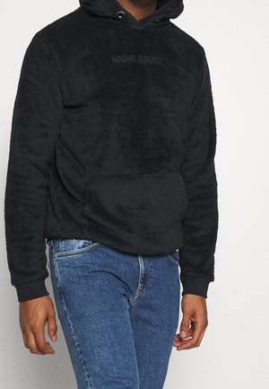 Personne portant un sweat à capuche noir et duveteux avec le texte "NIGHT ADDICT" et un jean bleu, debout devant un fond clair uni.