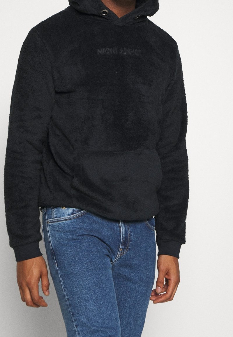 Personne portant un sweat à capuche noir et duveteux avec le texte "NIGHT ADDICT" et un jean bleu, debout devant un fond clair uni.