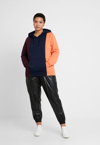 Sudadera con capucha en bloques de color naranja, azul marino y burdeos, con un bolsillo frontal. Combinada con pantalones jogger negros y brillantes y zapatillas blancas.
