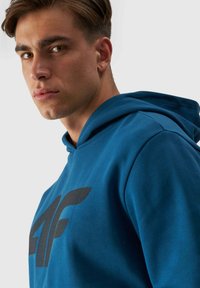 Sudadera azul con capucha, con un gran logo negro. La tela parece suave con una textura lisa y un diseño de corte relajado.