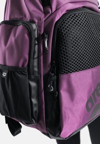 Zaino viola e nero con tessuto strutturato, dettagli in rete, molte tasche con cerniera e un logo audace sul lato.