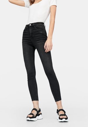 Femme portant un jean skinny noir taille haute, un top blanc à manches courtes et des sandales noires avec des semelles blanches épaisses, debout devant un fond uni.