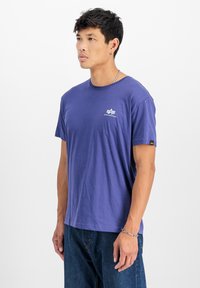 Violeta kokvilnas T-krekls ar apaļu izgriezumu, īsām piedurknēm un mazu baltu logo uz krūtīm. Uz piedurknes ir piekabe.