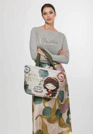 Frau mit zurückgebundenem dunklem Haar trägt ein graues Langarmshirt und einen bunten Rock, hält eine Tragetasche mit einem Cartoon-Mädchen und dem Text "Know Yourself".