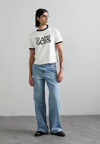 Bela skrajšana majica s črnim potiskom "TI AMO GCDS" in črnim rebrastim ovratnikom, v kombinaciji s svetlo modrimi širokimi jeans hlačami in črnimi čevlji.
