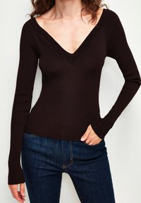 Top marron côtelé à manches longues avec un col en V profond. Caractérisé par une coupe ajustée et un tissu texturé. Associé à un jean bleu foncé.