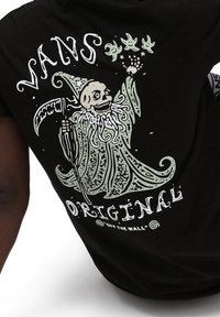 Camiseta negra con un gran gráfico caprichoso en verde y blanco de un esqueleto con una guadaña y elementos de diseño en espiral, de la marca "Vans."