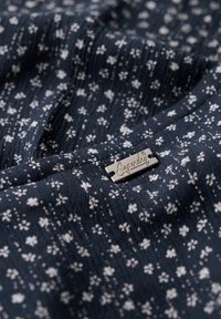 Granatowy materiał z teksturowaną powierzchnią, na którym znajdują się małe białe kwiatowe nadruki. Do tkaniny przymocowana jest srebrna metka z napisem "Superdry".