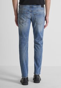 Svijetloplave traperice ravnog kroja, s dva stražnja džepa i blagim izbljeđenjem. Uparene s crnim cipelama, prikazane odostraga.