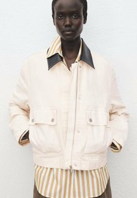 Cream-colored jacket with a black collar, front zip, and two buttoned pockets, layered over a tan and white striped shirt.