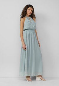 Helles blaues Maxikleid aus fließendem Stoff mit Halfterkragen, gerafftem Bauch und einem langen, locker fallenden Rock. Getragen mit weißen Sandalen.