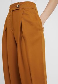 Rostfärgade högmidjade byxor med knappstängning och framfickor, bärs med en hand i sidfickan.