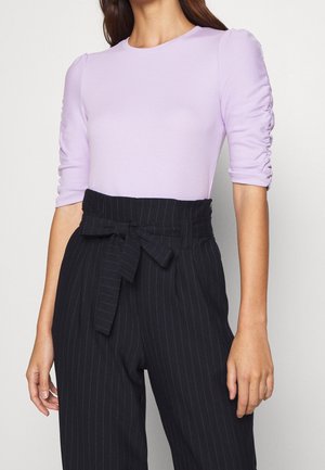 Mujer con una blusa de manga fruncida color púrpura claro, metida en unos pantalones de rayas negras de cintura alta con cinturón anudado en la cintura.