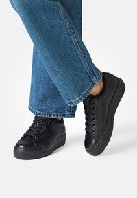 Schwarze Leder-Sneaker mit strukturierter Oberfläche, flacher Sohle und rundem Zehenbereich. Ausgestattet mit schwarzen Schnürsenkeln und einem weichen Innenfutter, kombiniert mit blauen Jeans.