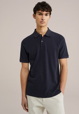 Polo din bumbac de culoare bleumarin, având guler și plachetă cu trei butoane. Mâneci scurte și o croială lejeră, purtat cu pantaloni de culoare deschisă.