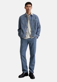 Denim jas in lichtblauw, met twee voorkruiszakken en knoopsluiting. Gelaagd met een geruit shirt, gecombineerd met bijpassende blauwe broek en zwarte schoenen.